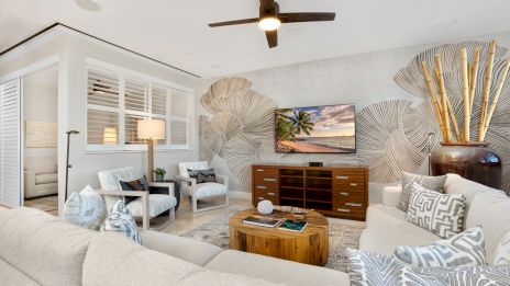 A bright, airy living room with light gray walls, tropical palm leaf wall decor, a wooden TV console, beige sofas, patterned throw pillows, and a ceiling fan, plus a warm coastal vibe.
