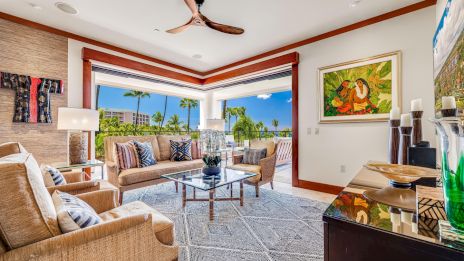 A bright tropical living room with beige woven sofas, a glass coffee table, large windows showing palm trees and blue skies, and a ceiling fan, ending this sentence.