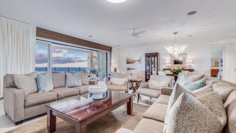 A bright, elegant living room with beige sofas, a wooden coffee table, large windows overlooking the ocean, and a chandelier in the dining area.