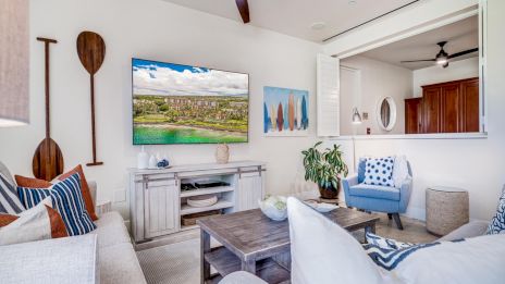 A bright beachy living room with a mounted TV, coastal decor, blue and white cushions, wicker baskets, and a gray wood coffee table, open to a kitchen. End.