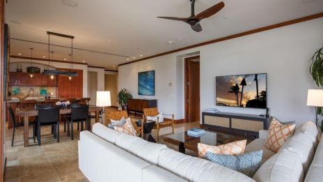 A modern open-plan living area with a white sofa, TV, wood accents, kitchen/dining in the background, warm lighting, and ceiling fan.