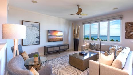 A bright, modern living room with a beige sectional, gray chairs, glass coffee table, and a TV on a slim stand; large windows offer a seaside view.