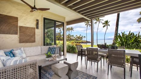 Outdoor tropical patio with wicker seating, patterned cushions, and a dining table overlooking palm trees and the ocean; modern ceiling fan above.