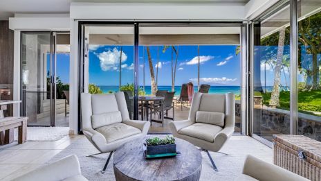 A bright coastal living room with cream lounge chairs, a round wooden coffee table, sliding glass doors, and an outdoor patio overlooking palm trees and the sea, perfectly cozy.