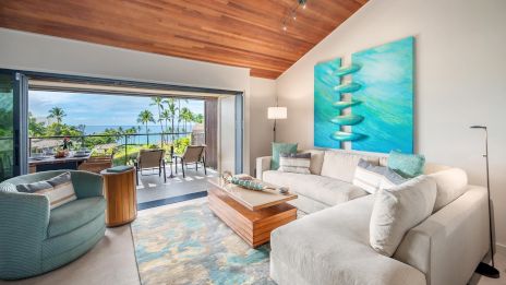 A bright, modern living room with a beige sectional, blue artwork, and a view opening to a sunny balcony and palm trees outside.