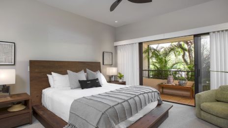 A modern bedroom with a low wooden bed, white linens, gray throw, matching nightstands, and a ceiling fan; sliding glass doors open to a tropical balcony.