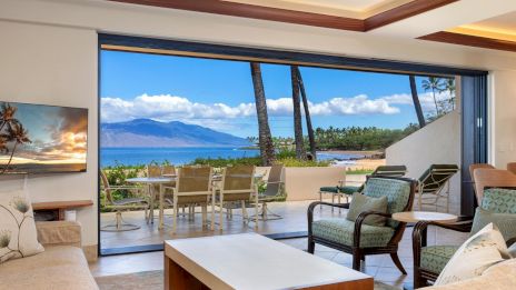 A bright tropical living room opens to a sunny terrace with palm trees, a dining table, and ocean view beyond the sliding glass doors.