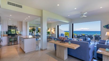 Bright open-concept coastal home: airy kitchen, laundry, and living areas flow to a shaded deck with blue couches and ocean view, sunset-ready.
