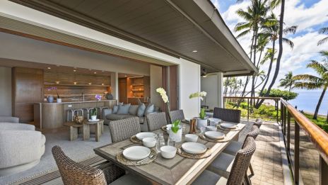 A stylish beachfront balcony with a long dining table set for a meal, wicker chairs, tropical plants, and a view of palm trees and the ocean.