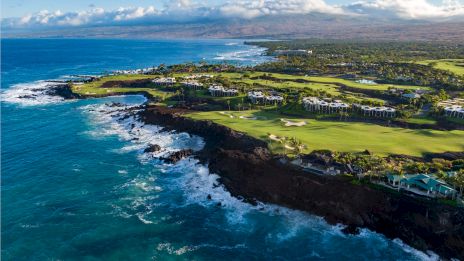 A dramatic coastline with rugged cliffs, turquoise waves, and a green golf course overlooking the ocean, sunny skies, and distant mountains.