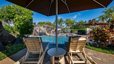 A sunny poolside scene with two lounge chairs under a large umbrella, a small table between them, overlooking a tranquil blue pool and tropical landscaping.