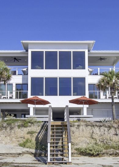 A modern beachfront house with large glass windows, palm trees, and a wooden stairway leading to the sandy yard, under a clear blue sky.
