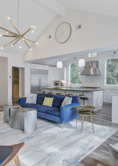 A bright, open-concept living area with a white kitchen, blue sofa, modern chairs, and a neutral rug and decor, seamlessly designed.