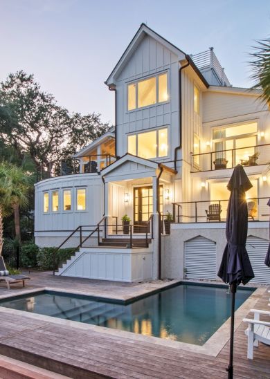 Luxurious modern house with a rectangular pool, wooden deck, palm trees, and outdoor lounge chairs under umbrellas.