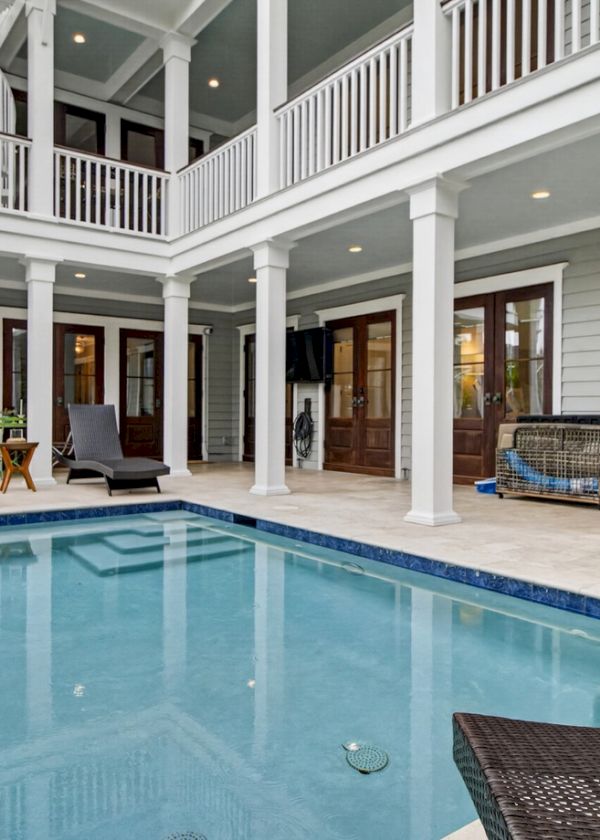 A modern house with a rectangular pool, white columns, covered patio, lounge chairs, and an American flag by the poolside.