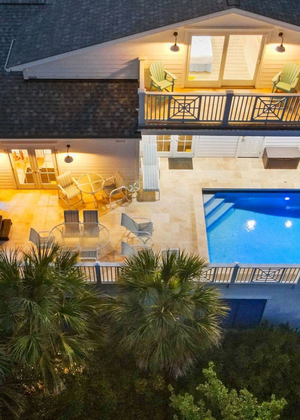 Aerial view of a two-story house with a bright rooftop balcony, a large rectangular pool and a smaller spa, surrounded by palm trees at dusk.