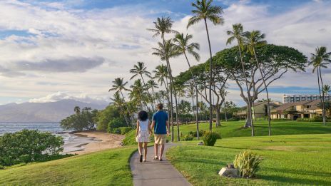 A couple walks along a seaside path lined with palm trees, grassy lawns, and a coastal view under a blue sky.