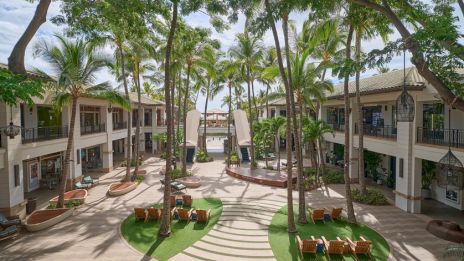 A sunny hotel courtyard with palm trees, circular green patches, curved steps, and patio seating between two-story beige buildings.
