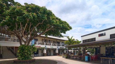 A sunny outdoor shopping plaza with a large tree, small storefronts, and seating areas, creating a relaxing open-air mall vibe.
