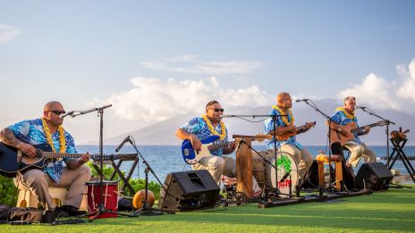 A four-member band sits on a grassy stage by the sea, playing guitars and drums with microphones, under a sunny, partly cloudy sky.