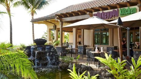 A sunny outdoor cafe with tropical plants, a water feature, and seating under umbrellas; sign says &ldquo;Monkeypuff kitchen.&rdquo;