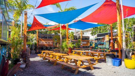 A sunny outdoor beer garden with picnic tables, colorful shade sails, tropical plants, and food trucks in a relaxed, festive setting.