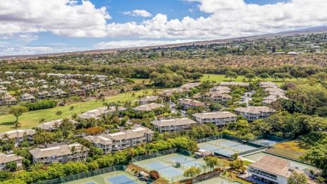 Aerial view of a residential area with rows of houses, tennis courts, and greenery under a partly cloudy sky, expansive suburb landscape.