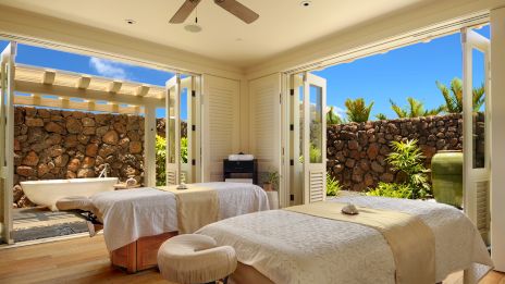 A bright spa treatment room with three massage beds, open doors to a stone courtyard, warm wood floors, and a ceiling fan.