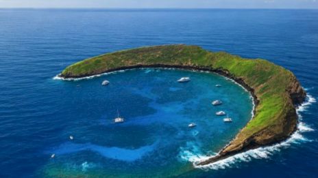 A crescent-shaped island with green cliffs forming a turquoise inner lagoon, surrounded by deep blue sea and several anchored boats, sunny skies.