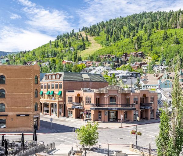 A small town with brick and stucco buildings sits at the base of green hills; a quiet street and trees frame the scene.