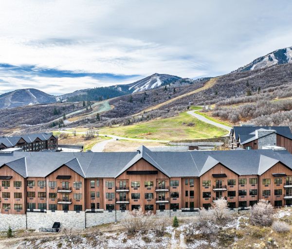 A row of brown brick apartment buildings with gray roofs sits at the base of rolling hills and light snow, winding trails on the slopes.