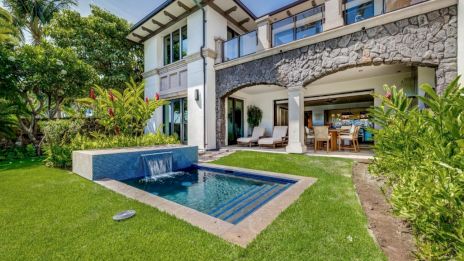 A luxurious two-story villa with a stone fa&ccedil;ade, lush tropical garden, a small pool with a spillover, and an open-air seating area under a covered patio, perfect for outdoor entertaining.
