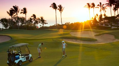 Two golfers on a sunny course near palm trees, a golf cart nearby, sand traps, and a bright sunset glow.