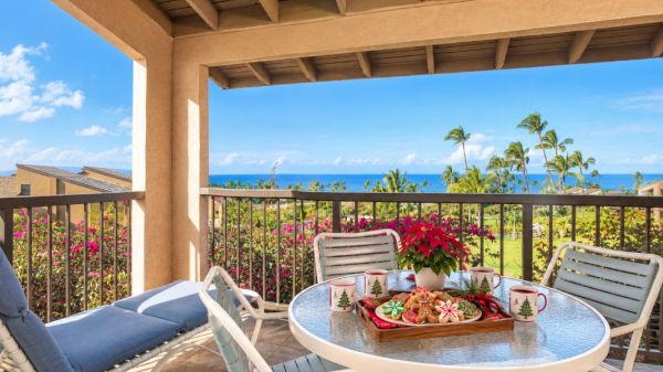 A balcony scene with a round glass table, four white chairs, red flowers centerpiece, and a view of palm trees and the blue ocean beyond, sunny and inviting.