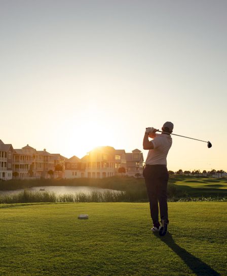 A person swings a golf club on a green fairway with houses and a sunset backdrop in the distance, creating a calm, sunlit scene.