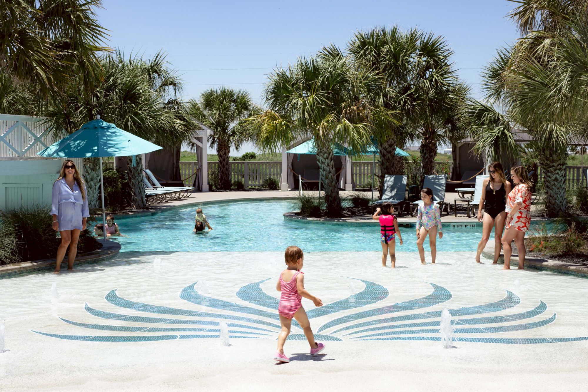 A sunny pool scene with kids playing, palm trees, lounge chairs, and umbrellas; a little girl in pink walks across a decorative blue pattern by the water.