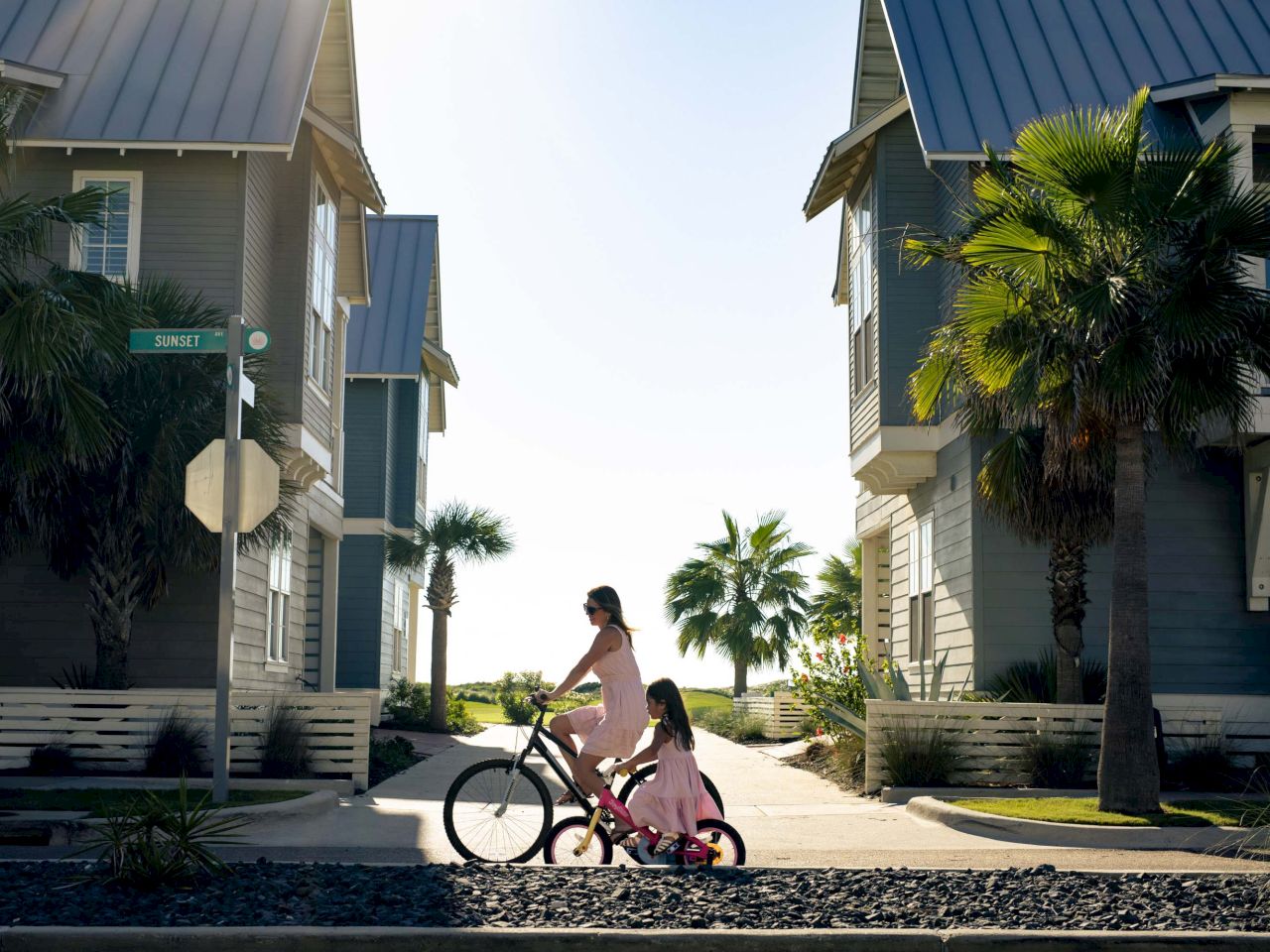 A person on a pink bicycle with a child seat is riding between two blue beach houses near palm trees by the water, sunny and bright.