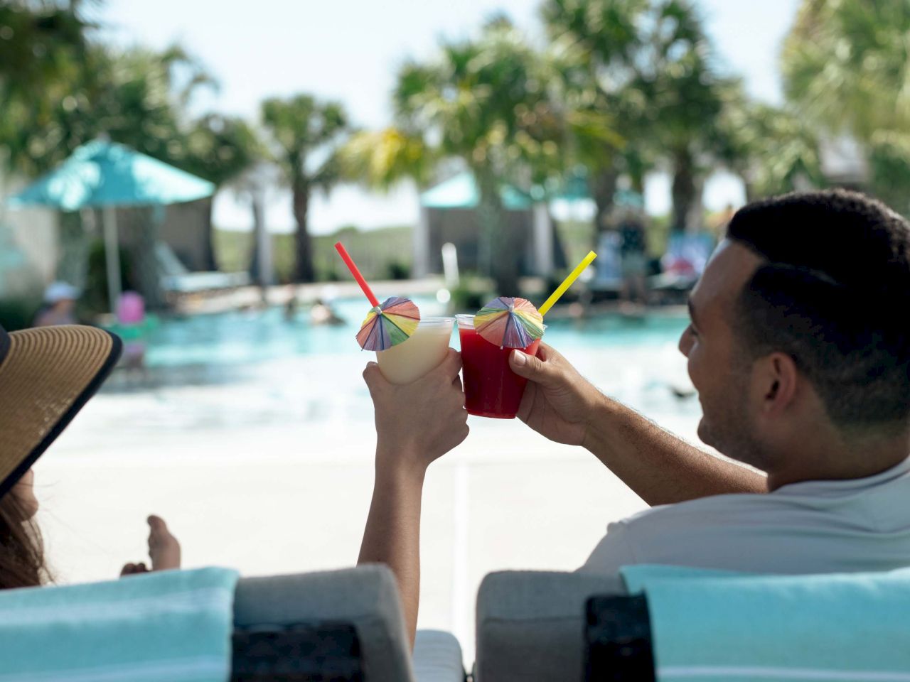 Two people lounging poolside, toasting colorful cocktails with umbrellas under sunny, palm-tree scenery.
