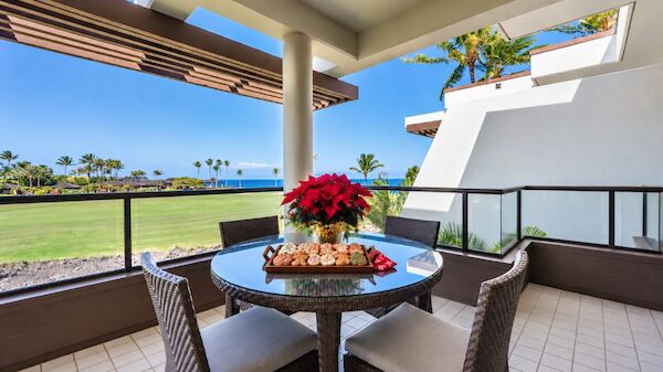 A sunny balcony with a round glass table and four wicker chairs, red poinsettia centerpiece, overlooking a green lawn and distant palm trees.