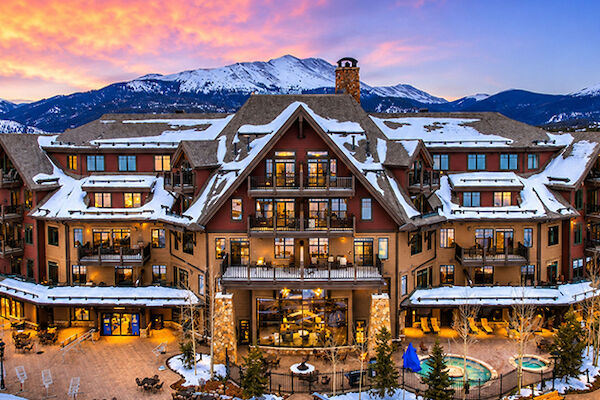 A grand alpine resort with snow-covered roofs, warm windows, and a central courtyard, set against snowy mountains at sunset, with outdoor pools and seating.