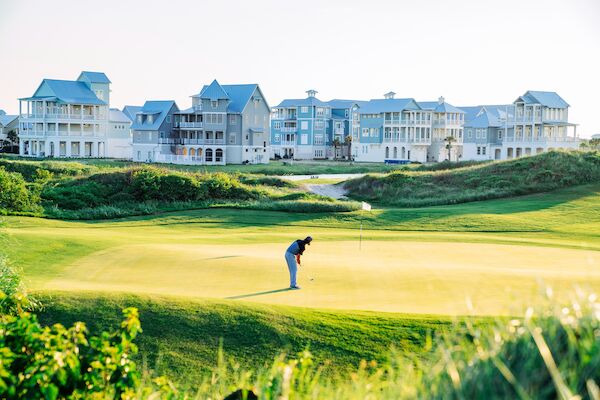 A golfer on a sunny green beside a row of seaside townhouses or resort buildings, with lush fairways and sand, peaceful and bright.