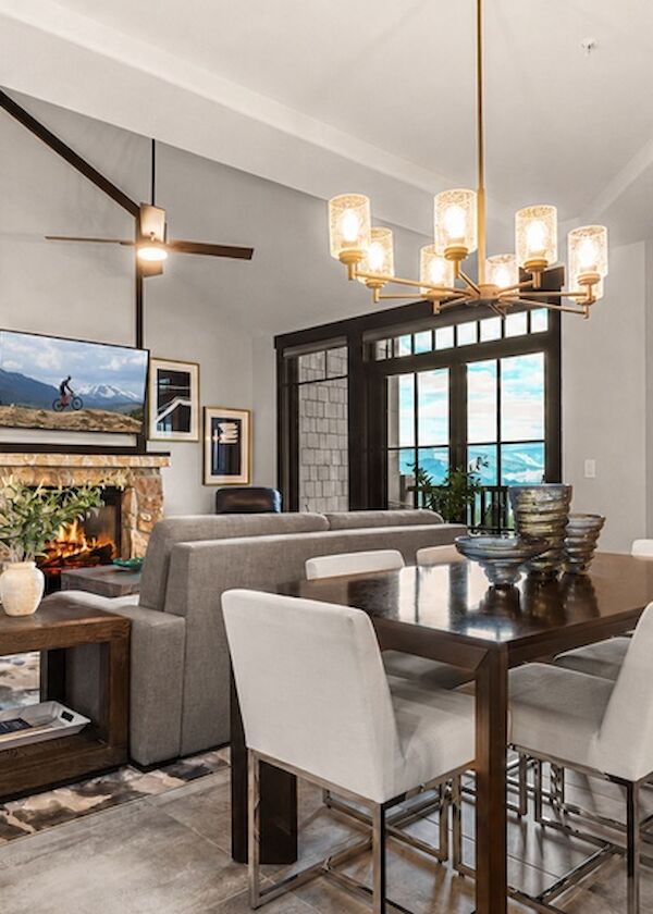 Modern living-dining room with a cozy fireplace, gray sectional, farmhouse dining table, and white upholstered chairs under a chandelier.