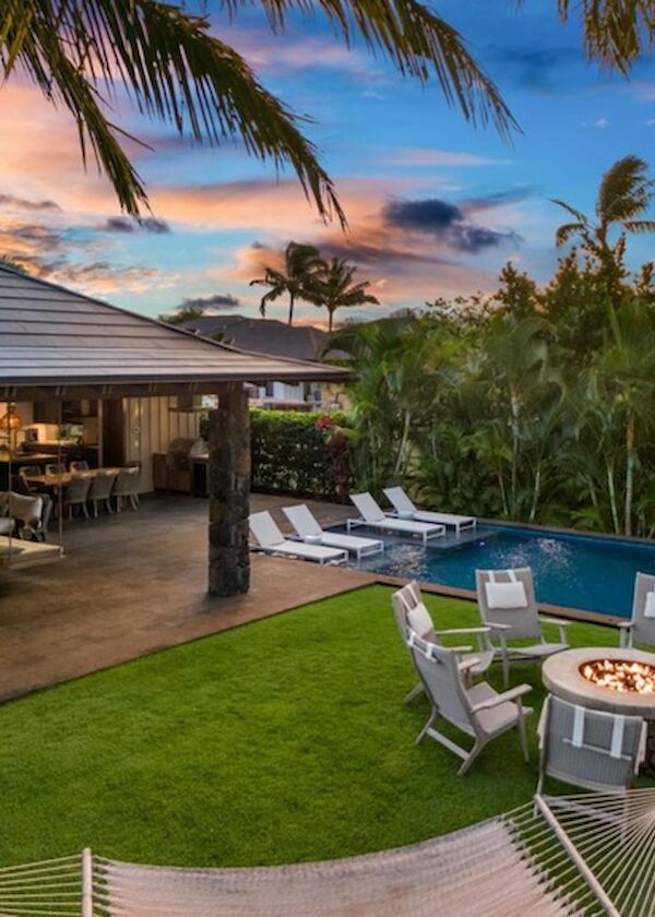 A tropical poolside villa with lounge chairs, a fire pit table, hammock, palm trees, and an open-air seating area at sunset.