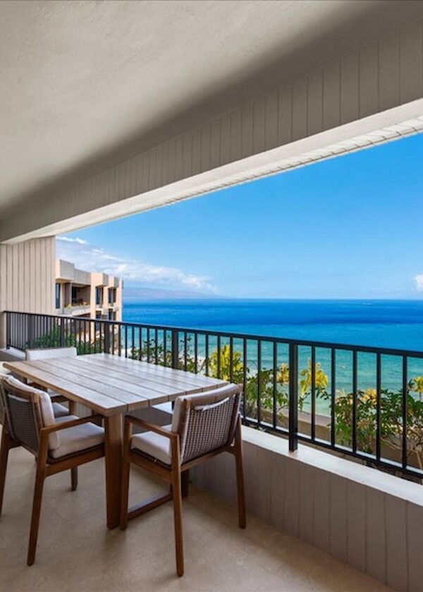 A spacious balcony with a wooden dining table and chairs overlooks a blue ocean and clear sky, offering a coastal view.