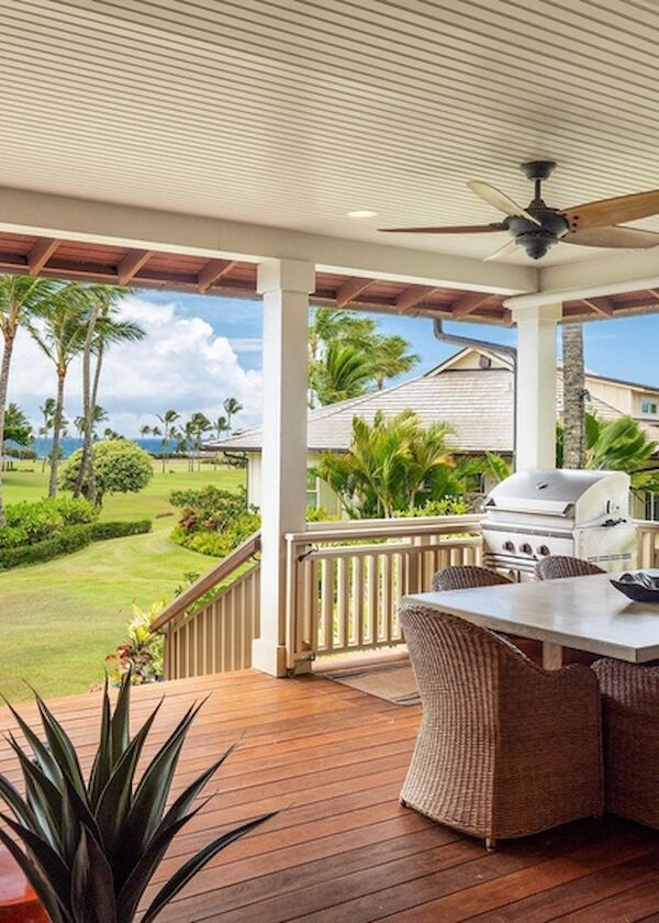A covered outdoor patio with a wooden deck, wicker dining set, ceiling fans, and a grill, overlooking a sunny, tropical lawn and palm trees.