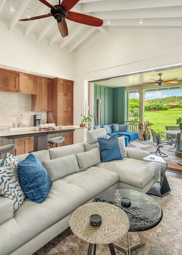 A modern, open-concept living room and kitchen with a light gray sectional, blue accent pillows, wood cabinets, and a breezy sliding door to a sunny outdoor space.