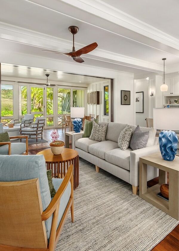 A bright, open-concept living room with a white sofa, blue accent pillows, wooden chairs, a cozy rug, and a modern kitchen in the background, airy and inviting.