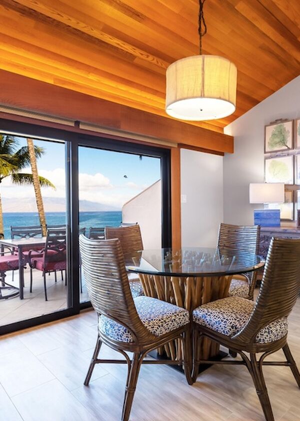 A sunny tropical dining area with a glass door opening to a seaside patio, palm trees, and a cozy wooden ceiling, inviting ocean views.