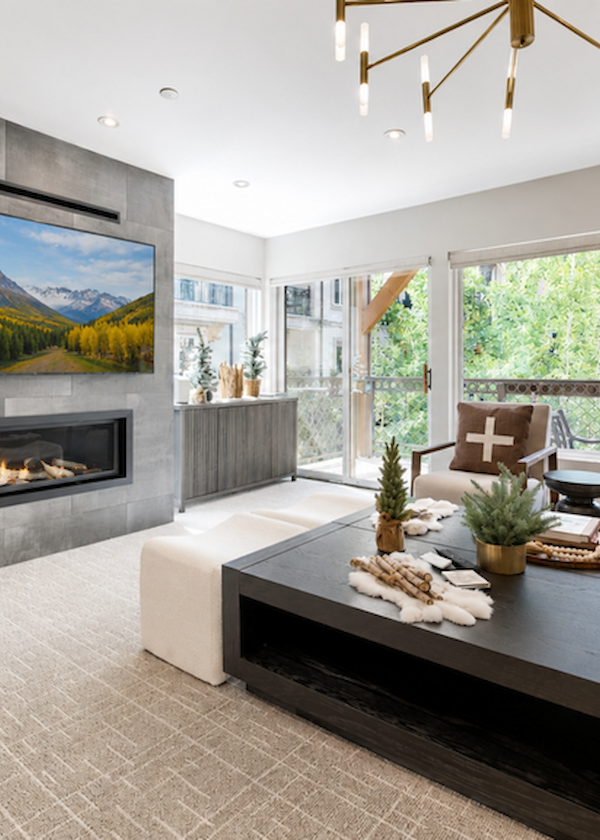 A modern living room with a wall-mounted fireplace, a large TV, cozy seating, pine decor, and big windows offering natural light.