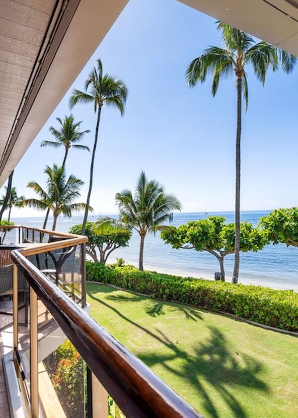 A sunny beachfront balcony with a wooden railing, palm trees, lush green lawn, and a clear blue ocean view.